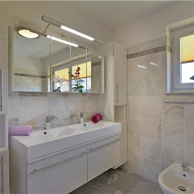 A modern bathroom with bright, elegant design. There is a double sink, a large mirror, and a plant on the windowsill.