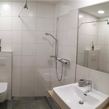 A modern bathroom with a shower, a sink, and a toilet. The walls are clad with white tiles and there is a large mirror.