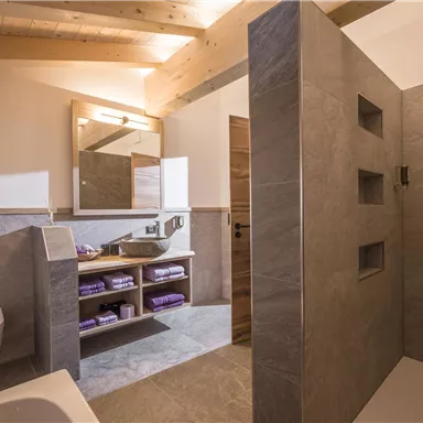 Ein modernes Badezimmer mit einer Dusche und einer Badewanne. Die Wände sind mit grauen Fliesen verkleidet und es gibt eine große Spiegelfläche.