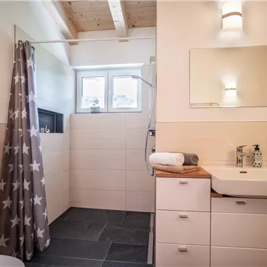 Ein modernes Badezimmer mit einer Dusche und einem Waschbecken. Die Wände sind hell gefliest und es gibt einen großen Spiegel.