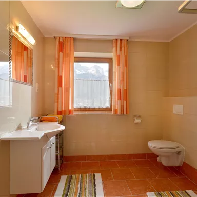 Ein modernes Badezimmer mit orangefarbenen Vorhängen und einem großen Fenster. Es gibt eine Dusche, ein WC und eine Waschbeckenanlage.