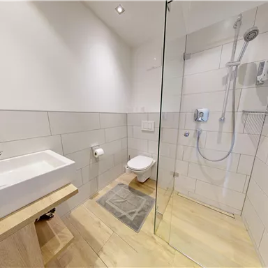 A modern bathroom with a shower, sink, and toilet. The walls are light tiled and the floor is made of wood.