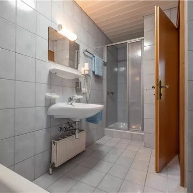 A modern bathroom with gray tiles. There is a shower, a sink, and a mirror as well as towels on a shelf.