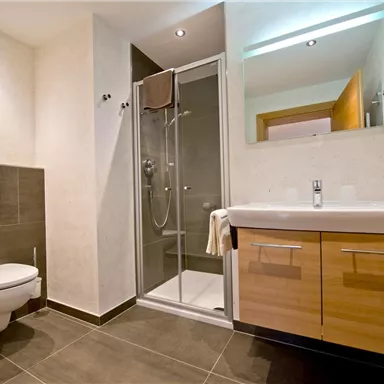 Ein modernes Badezimmer mit einer Dusche und einem Waschbecken. Die Wände sind hell und der Boden ist dunkel gefliest.