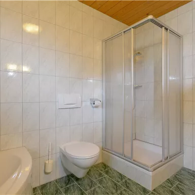 A bright bathroom with a shower and a bathtub. The walls are tiled and the floor has a green covering.