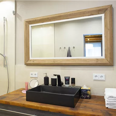 Ein modernes Badezimmer mit einer großen Spiegelfläche und einem eleganten Waschbecken aus schwarzem Material. Die Holzoberfläche und die Duschkabine verleihen dem Raum eine einladende Atmosphäre.