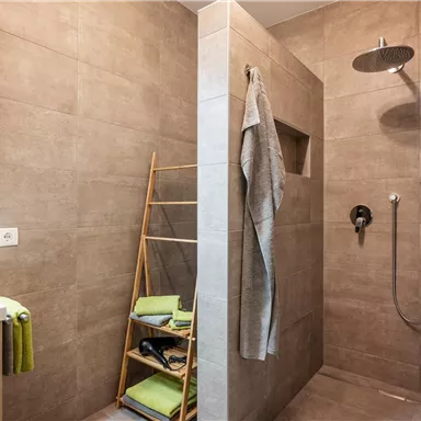 Moderne Dusche mit grauen Fliesen und einem rechtwinkligen Duschbereich. Ein Handtuchständer und Pflegeartikel sind im Raum sichtbar.