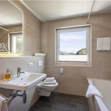 A modern bathroom with a bathtub and a sink. Sunlight streams in through the window and there are fresh towels.