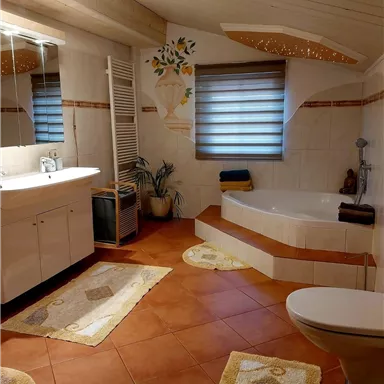 Ein helles Badezimmer mit modernen Fliesen und einer Badewanne. Die Einrichtung ist schlicht und einladend, mit Pflanzen und dekorativen Matten.
