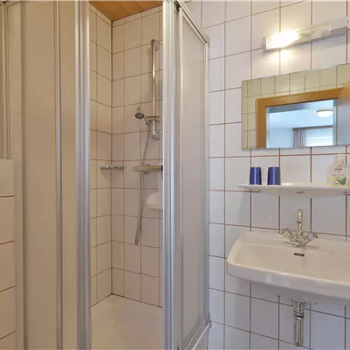 A simple bathroom with a shower, a sink, and a mirror. The walls are tiled and there are basic toiletries.