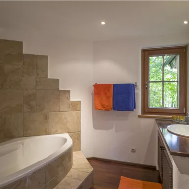 Ein modernes Badezimmer mit einer Badewanne und einer Dusche. Bunte Handtücher hängen an der Wand und ein Fenster sorgt für Tageslicht.
