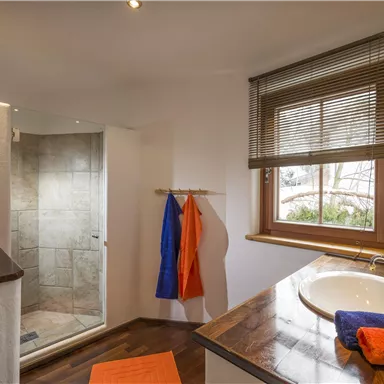 Ein helles Badezimmer mit einer Dusche und einem Waschbecken. Bunte Handtücher hängen an der Wand und ein Fenster sorgt für natürliches Licht.