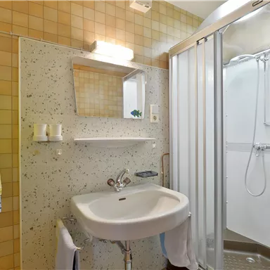 A small bathroom with a shower, a sink, and a mirror. The walls are covered with light tiles in a minimalist design.