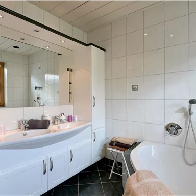 A modern bathroom with a bathtub and a large mirror. The walls are tiled and the fixtures are in white.