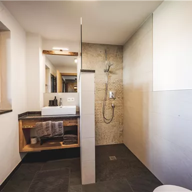 Ein modernes Badezimmer mit einer offenen Dusche und einer Holzablage. Helle Wände und große Fenster sorgen für eine freundliche Atmosphäre.