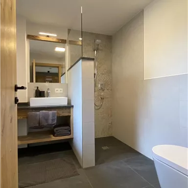 Ein modernes Badezimmer mit grauen Fliesen und einem eleganten Waschtisch. Die Dusche ist durch eine Glaswand abgetrennt.