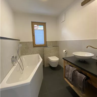 Ein modernes Badezimmer mit einer Badewanne und einem Waschbecken. Die Wände sind in einem neutralen Farbton gehalten und es gibt ein Fenster für natürliches Licht.
