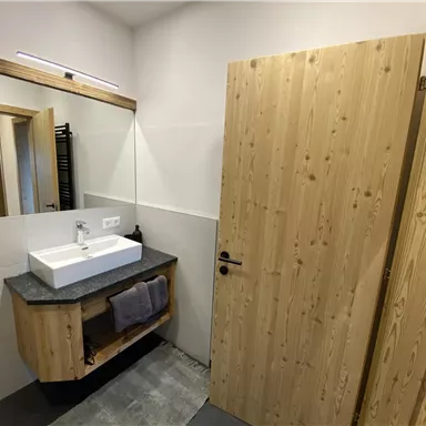 Ein modernes Badezimmer mit Holzverkleidung und einem Waschbecken auf einer grauen Arbeitsplatte. Im Hintergrund befindet sich ein Spiegel und eine offene Tür, die zu einem anderen Raum führt.