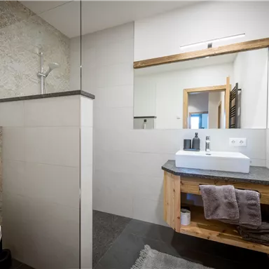Ein modernes Badezimmer mit einer begehbaren Dusche und einer eleganten Waschgelegenheit. Holz- und Steinakzente verleihen dem Raum eine warme Atmosphäre.