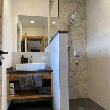 Ein modernes Badezimmer mit einer Dusche und einem Waschbecken. Die Wände sind hell gefliest und es gibt Holzregale für Handtücher.