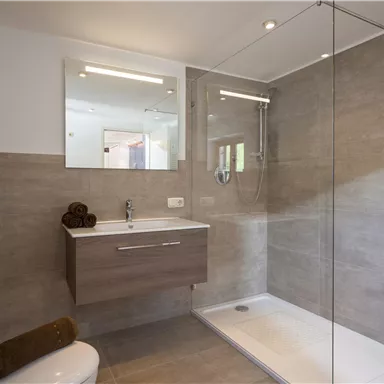 A modern bathroom with a glass shower and light tiles. The amenities include a sink with a mirror and towels.