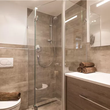 A modern bathroom with a glass shower and neutral colors. Towels are neatly arranged on the shelf, and a plant decorates the space.
