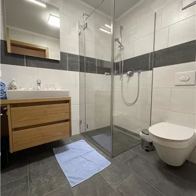 A modern bathroom with a glass shower and a wooden dresser. The floor is tiled with dark tiles and a blue bath mat is on the floor.