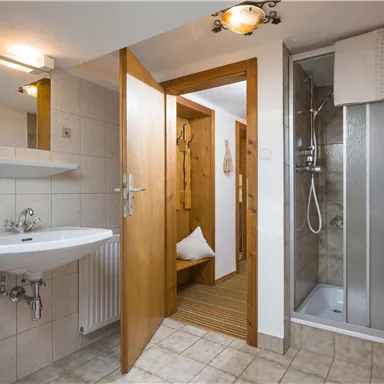 A modern bathroom with a shower, sink, and wooden details. The walls are designed with light tiles, and there is an open door to another room.