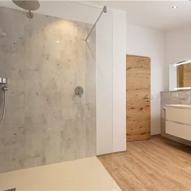 A modern bathroom with a walk-in shower and a light wooden floor. The space is minimalist and well-lit, featuring a large mirror and sleek furniture.