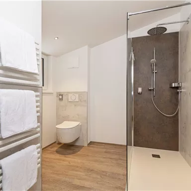 A modern bathroom with a shower and a toilet. Towels hang on a radiator, and the floor is designed with a wood look.