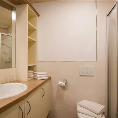 Ein modernes Badezimmer mit einer Dusche und einem Waschbecken. Es gibt helle Wände und Holzregale mit frischen Handtüchern.