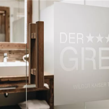 An elegant bathroom with a modern sink and paneled wood design. The glass door features the inscription “DER GREIL” in stylish design.