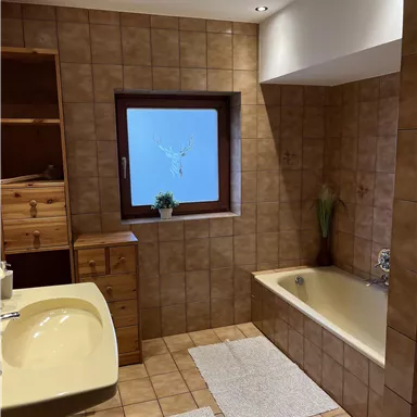 Ein Badezimmer mit braunen Fliesen und einer Badewanne. Es gibt ein Fenster, einen Holzschrank und zwei Teppiche auf dem Boden.