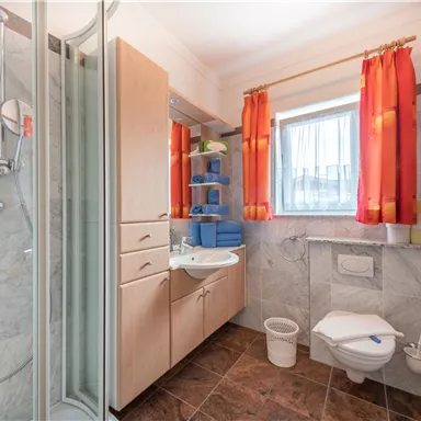 A bright bathroom with a shower, sink, and toilet. The windows have orange curtains and shelves with blue containers.