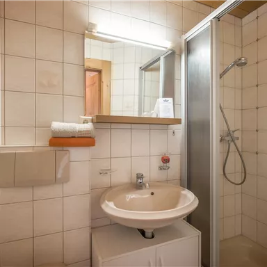 Ein kleines Badezimmer mit einer Dusche und einem Waschbecken. Die Wände sind gefliest und es gibt einige Handtücher auf einem Regal.