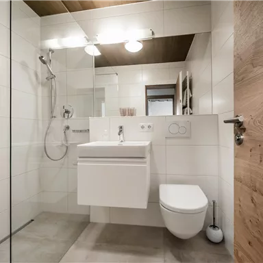 A modern bathroom with a shower and a sink. The walls are light-colored tiled and there are wooden details.