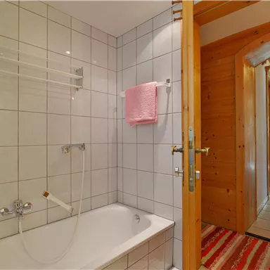 A bright bathroom with a bathtub and white tiles. A hallway is visible in the background with wooden details.