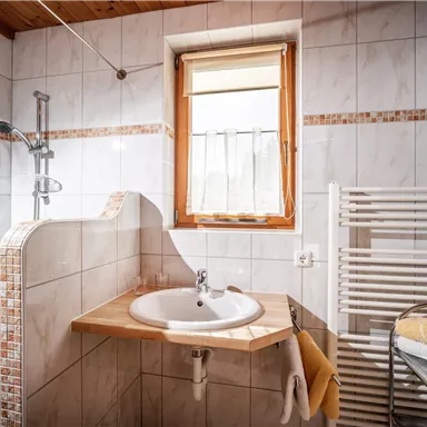 Ein modernes Bad mit weißen Fliesen und einem Waschbecken. Es gibt eine Dusche und einen Handtuchheizkörper sowie viel Tageslicht durch das Fenster.