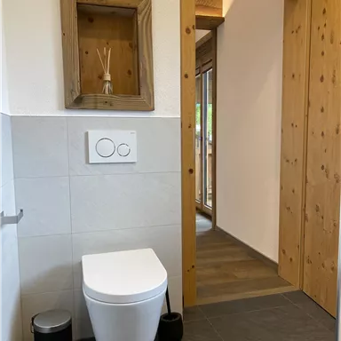 Ein modernes Badezimmer mit einer weißen Toilette und einem hellen Holztürrahmen. An der Wand ist ein Holzregal mit Duftstäbchen zu sehen.