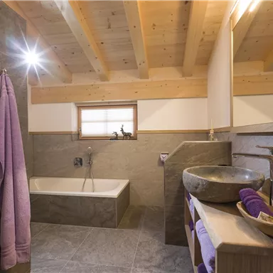 Ein modernes Badezimmer mit einer Badewanne und einer Steinwaschbecken. Die Wände sind teilweise mit einem eleganten grauen Material verkleidet, und es gibt handtücher in Lila.