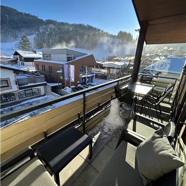 A balcony with seating and a view of a snow-covered village. In the background, mountains and a blue sky are visible.