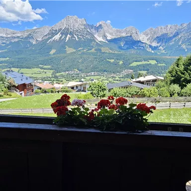 A picturesque mountain landscape with clear blue sky and green meadows. In the foreground, colorful flowers are blooming on a balcony.