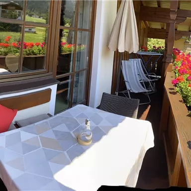 Ein gemütlicher Balkon mit einem Tisch und Stühlen, umgeben von bunten Blumen. Im Hintergrund sind grüne Wiesen und ein heller Himmel zu sehen.
