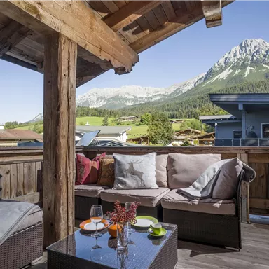 Ein gemütlicher Balkon mit einer Sitzgelegenheit und Blick auf majestätische Berge. Der klare Himmel und die Sonnenstrahlen schaffen eine einladende Atmosphäre.