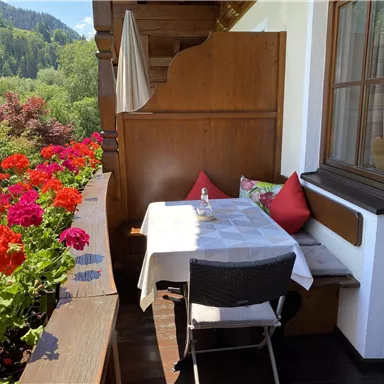 Ein gemütlicher Balkon mit einem Tisch und einer Bank. Bunte Blumen umgeben den Balkon und die Aussicht auf die Natur ist schön.