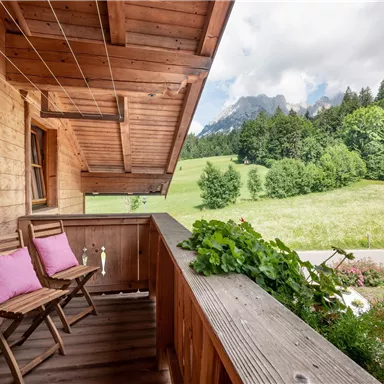 A cozy wooden balcony with two chairs and cushions. In the background, a green meadow and wooded mountains stretch out.