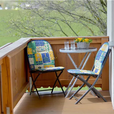 Ein kleiner Balkon mit zwei bunten Stühlen und einem Tisch. Auf dem Tisch stehen ein paar Gläser Wasser und eine Blumenschale.