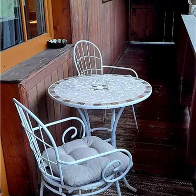 Ein kleiner Balkon mit einem runden Tisch und zwei weißen Stühlen. Die Holzverkleidung und der Boden verleihen eine gemütliche Atmosphäre.