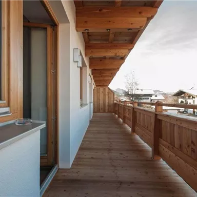 A spacious balcony with a wooden railing and a view of a mountainous landscape. The surroundings are bright and inviting, perfect for relaxing.
