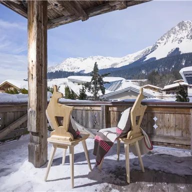 Ein Balkon mit zwei Stühlen aus Holz und einer Decke. Im Hintergrund sind schneebedeckte Berge und ein klarer Himmel zu sehen.